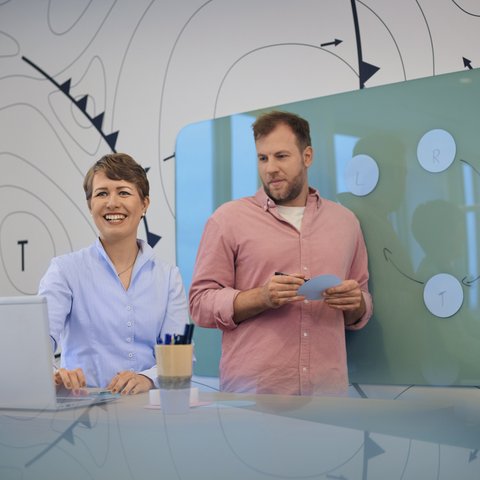 Ein Mann und eine Frau stehen vor einem grünen Whiteboard mit grafischen Elementen. Beide halten Karten in der Hand und unterhalten sich.
