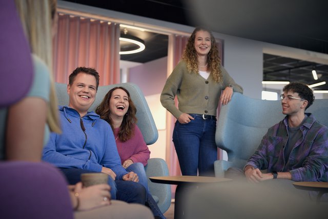 Dein Impact Projektmanagement Sechs Personen sitzen und stehen in einem Lounge-Bereich mit Sesseln. Sie unterhalten sich und lachen. Im Hintergrund sind Vorhänge und moderne Deckenleuchten zu sehen.