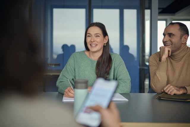 Unser Tech-Stack Softwareentwicklung & Testung Eine Frau spricht lächelnd, ein Mann sitzt daneben. Im Vordergrund ist eine Person mit Smartphone und Kaffeebecher unscharf zu sehen.