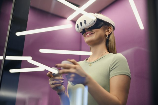 Unser Tech-Stack Projektmanagement Eine Frau trägt eine VR-Brille und hält Controller in den Händen. Sie steht vor einer lila Wand mit leuchtenden, geometrischen Lichtinstallationen.