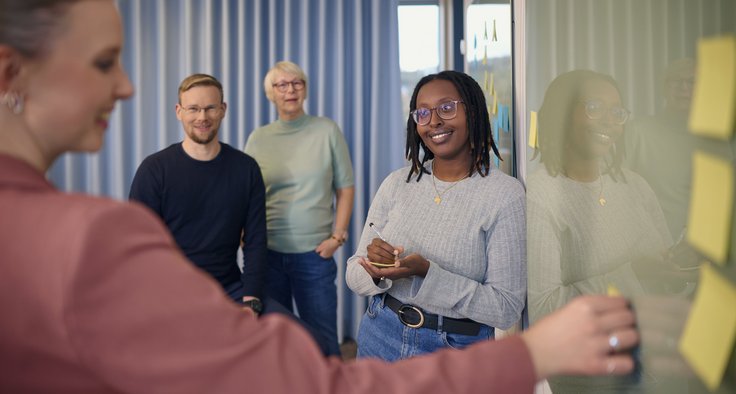 Vier Personen stehen vor einem Whiteboard mit Post-its. Eine Frau schreibt etwas auf, die anderen beobachten oder lachen.