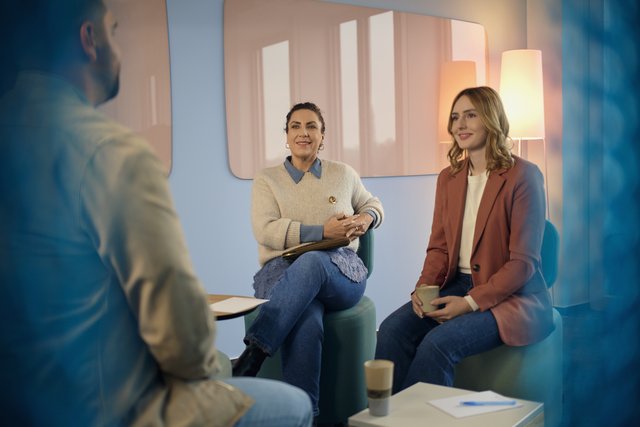 Dein Impact Consulting & IT-Beratung Zwei Frauen sitzen mit Getränken in einem Lounge-Bereich und sprechen mit einer weiteren Person im Vordergrund. Auf dem Tisch liegen Unterlagen und ein Becher.