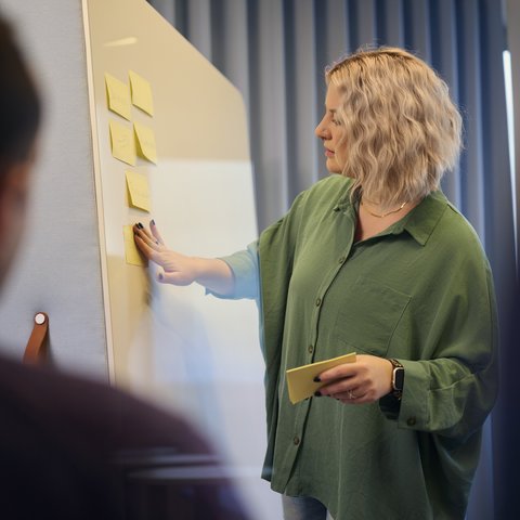 Eine Frau steht vor einem Whiteboard und arbeitet mit gelben Haftnotizen. Im Vordergrund sind andere Personen leicht unscharf zu sehen.