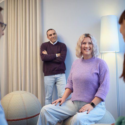 Vier Personen stehen oder sitzen in einem Raum mit heller Einrichtung. Eine Frau im Vordergrund lächelt, zwei weitere Personen sitzen, eine Person steht an der Wand.